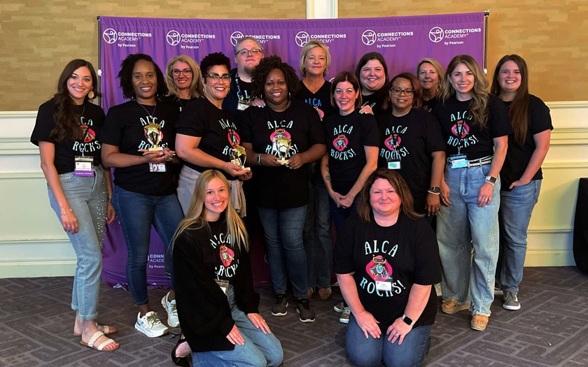 Group of fifteen diverse people in matching black t-shirts, smiling, standing and kneeling in front of a purple banner with logos.