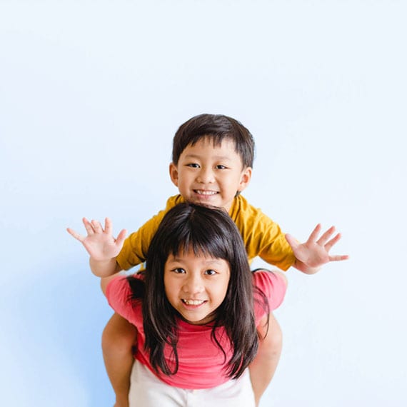 A young child in a yellow shirt sits on the shoulders of another young child in a pink top against a plain light blue background.