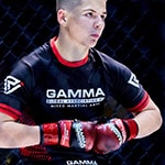 Ethan wearing a black and red GAMMA t-shirt and red gloves stands inside a caged fighting ring