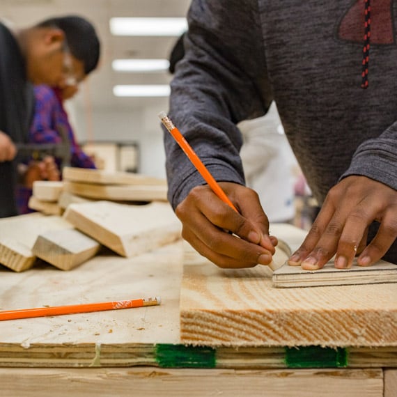 A person in a workshop environment using a pencil to mark measurements on a piece of wood. 
