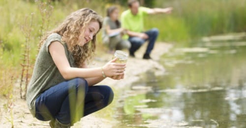 A student in a green shirt sitting near a lake collecting pond water for an experiement. 