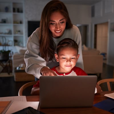 A Learning Coach mom in a white sweater and her young student in a red and grey shirt transferred mid-year to Connections Academy and are working on an online assignment on a laptop. 