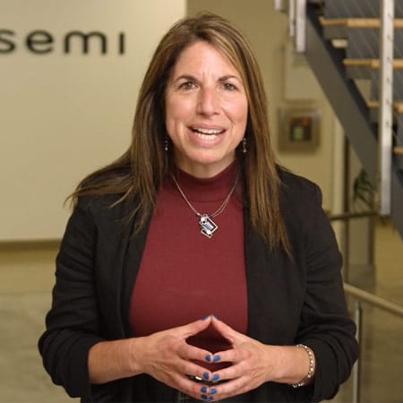 A person standing indoors, wearing a black blazer over a maroon top, with hands clasped.
